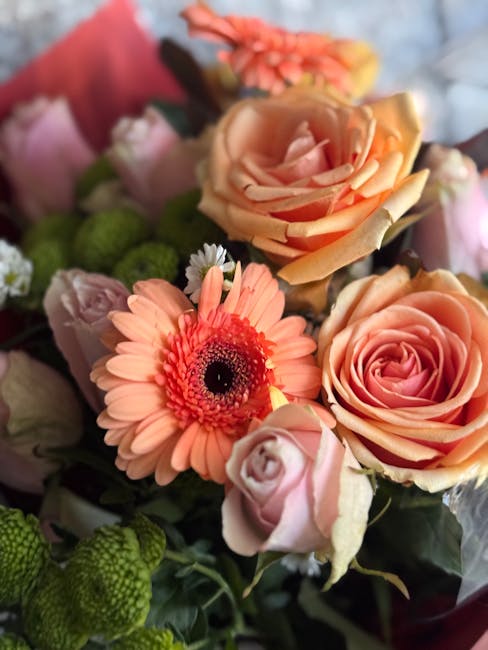 A close-up view of a floral arrangement featuring peach and pink roses, an orange-pink gerbera daisy, and small green chrysanthemums, all arranged with green foliage. The bouquet is wrapped with a pin