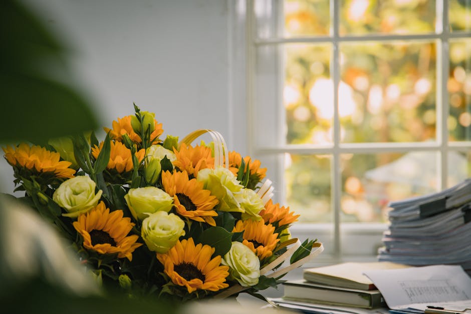 A vibrant floral arrangement featuring bright yellow sunflowers with dark centers and creamy white roses, placed on a cluttered desk next to neatly stacked documents and a yellow notepad. The bouquet 