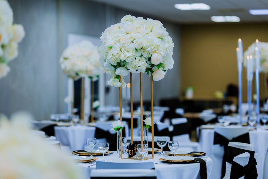 A floral arrangement featuring a large, spherical cluster of creamy white hydrangeas and roses, mounted on tall, slender gold stands with green ribbon accents, set on a dining table with white tablecl