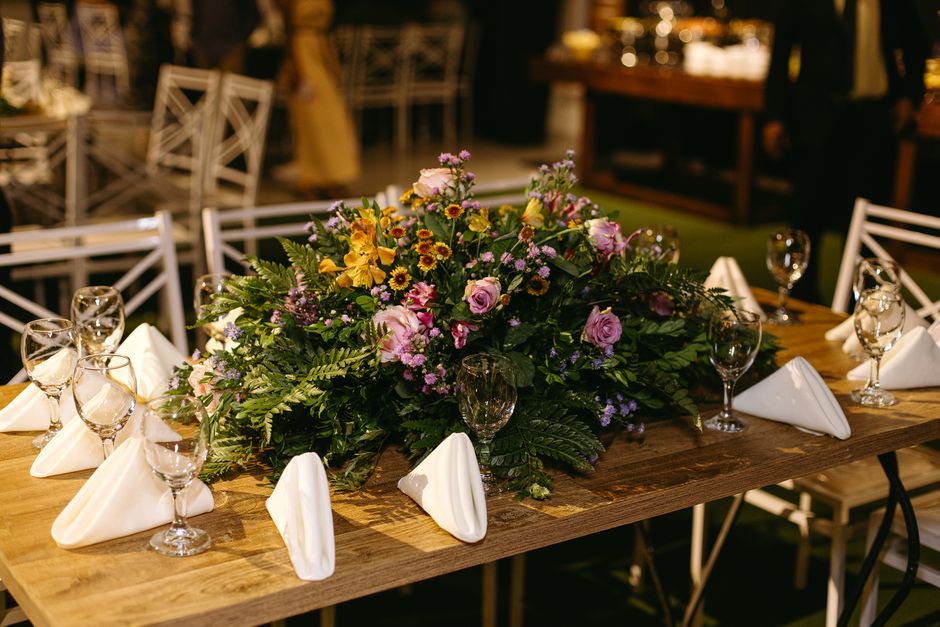 A large floral arrangement featuring pink roses, yellow daisies, and purple statice, accented with lush green ferns. The bouquet is placed on a wooden dining table set for an event, with neatly folded