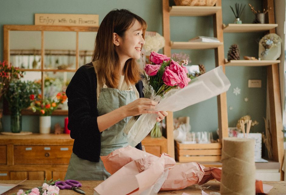 A smiling female florist, wearing a black long-sleeved top and a light grey apron, holds a bouquet of fresh pink roses and pink lisianthus wrapped in clear cellophane. She stands inside a cozy floral
