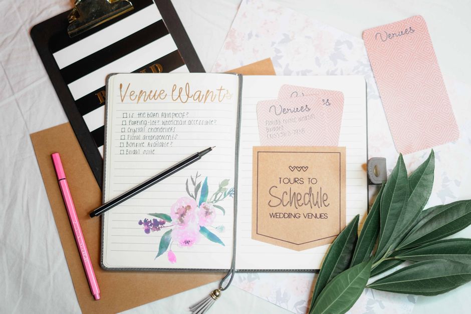 A light-colored table features an open notebook with a floral watercolor illustration of pink and purple flowers and green leaves on the page titled 'Venue Want,' next to a black pen. There are severa