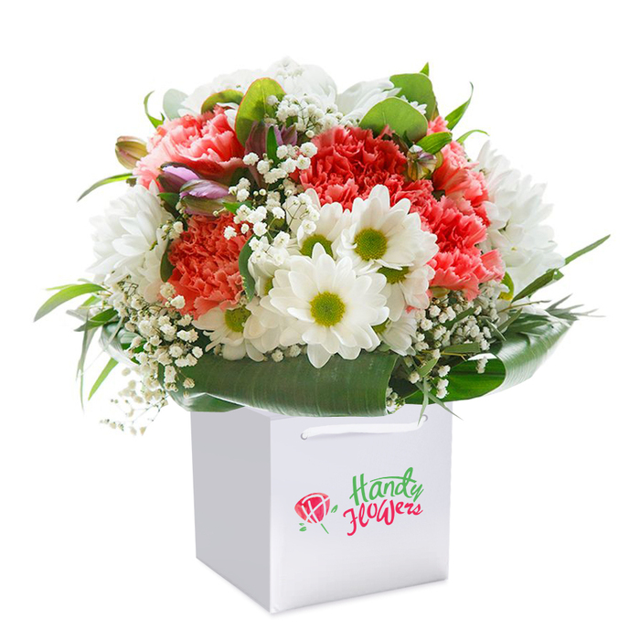 Within a crisp white gift bag, set against a pure white backdrop, a stunning bouquet reflects the heartfelt artistry of Harefield's finest florists. The arrangement draws the eye with its expertly placed blend of soft coral, vibrant peach, and deep red carnations-each bloom showcasing undulating, frilly petals full of texture and life, reminiscent of the colourful blooms in Harefield's beloved cottage gardens. Bright white daisy-like flowers are interspersed amongst the carnations, boasting lively yellow-lime centers that bring an invigorating, cheerful contrast, as if capturing the clear light that dances across the local fields. Wispy sprigs of dainty baby's breath are gently threaded throughout, softening the overall look and adding airiness and movement, while on the left, subtle buds touched by pale purple promise tomorrow's blossoms. The composition is encircled by luxuriant, glossy green leaves, providing a strong natural frame and a feeling of rooted abundance, mirroring the lushness of Harefield's landscape. Carefully shaped into a rounded, harmonious form, the bouquet exudes an inviting fullness ideal for celebratory moments-a birthday treat near Harefield's High Street or a heartfelt gesture sent to a neighbour. Sturdy ribbon handles on the bag make the arrangement perfect for effortless delivery across Harefield and its family-friendly avenues. Each detail is brought into sharp relief by the bright white background, emphasising the bouquet's crisp freshness and the pride that Harefield's florists bring to their craft-a tribute to the beauty and kindness at the heart of the local community.